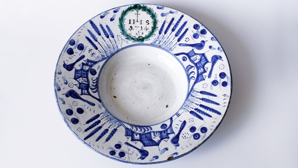 Fajánsový talíř, habánská keramika, tvaru kardinálského klobouku, na okraji kobaltem malovaná krajina s architekturami a ptáky, v zeleném věnci iniciály IHS a letopočet r. 1714. Zdroj: Národní muzeum