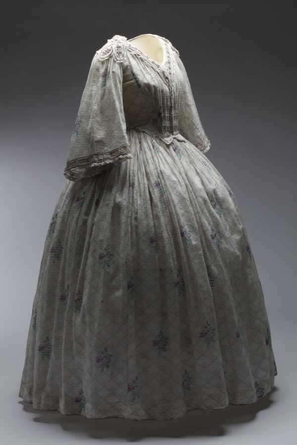 Šaty dámské bílé s drobným fialovým květinovým vzorem, tištěné jemné bavlněné plátno – mušelín, 1850–1860. Šaty pravděpodobně patřily principálce kočovné herecké společnosti Elišce Zöllnerové.
