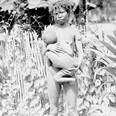 Stojící žena drží v náručí dítě (už dost velké), které kojí. Nová Guinea, 1896